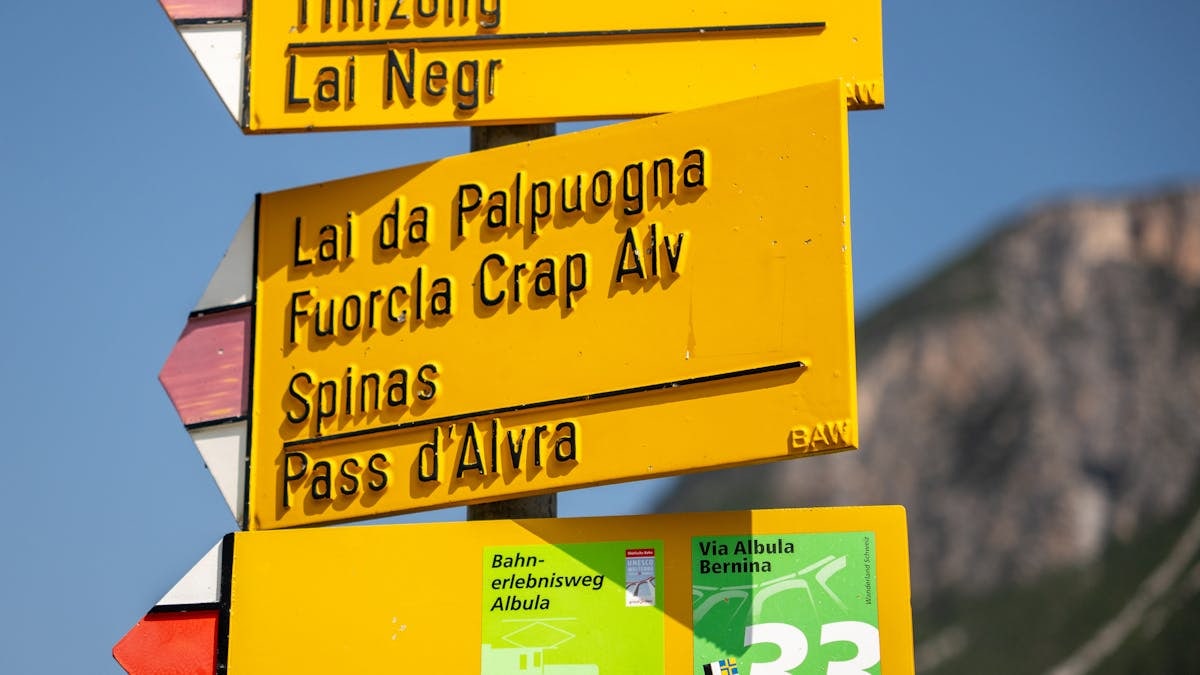 Close-up of a yellow trail sign in La Punt Chamues-ch, Switzerland, with mountains in the background.